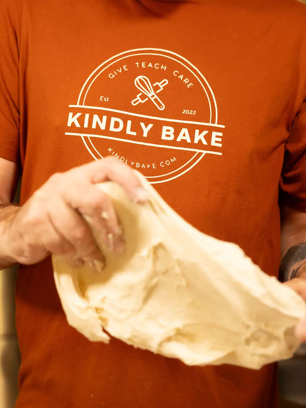 Close-up of a baker wearing a Kindly Bake branded orange t-shirt, stretching and folding a large piece of smooth, elastic sourdough dough by hand.