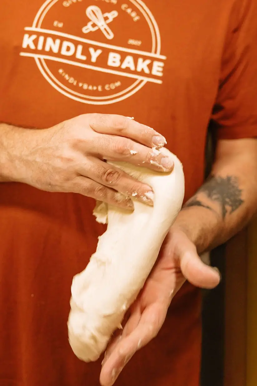 Kindly Bake owner, James Purcell, wearing a branded shirt and using both hands to gently round and shape a smooth, raw ball of sourdough dough.